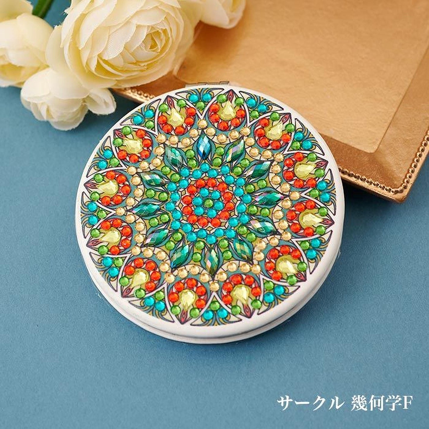A decorative round mirror with a colorful, intricate design featuring various shapes, patterns, and gemstones. The mirror is placed on a blue surface, with a bouquet of white flowers in the background.