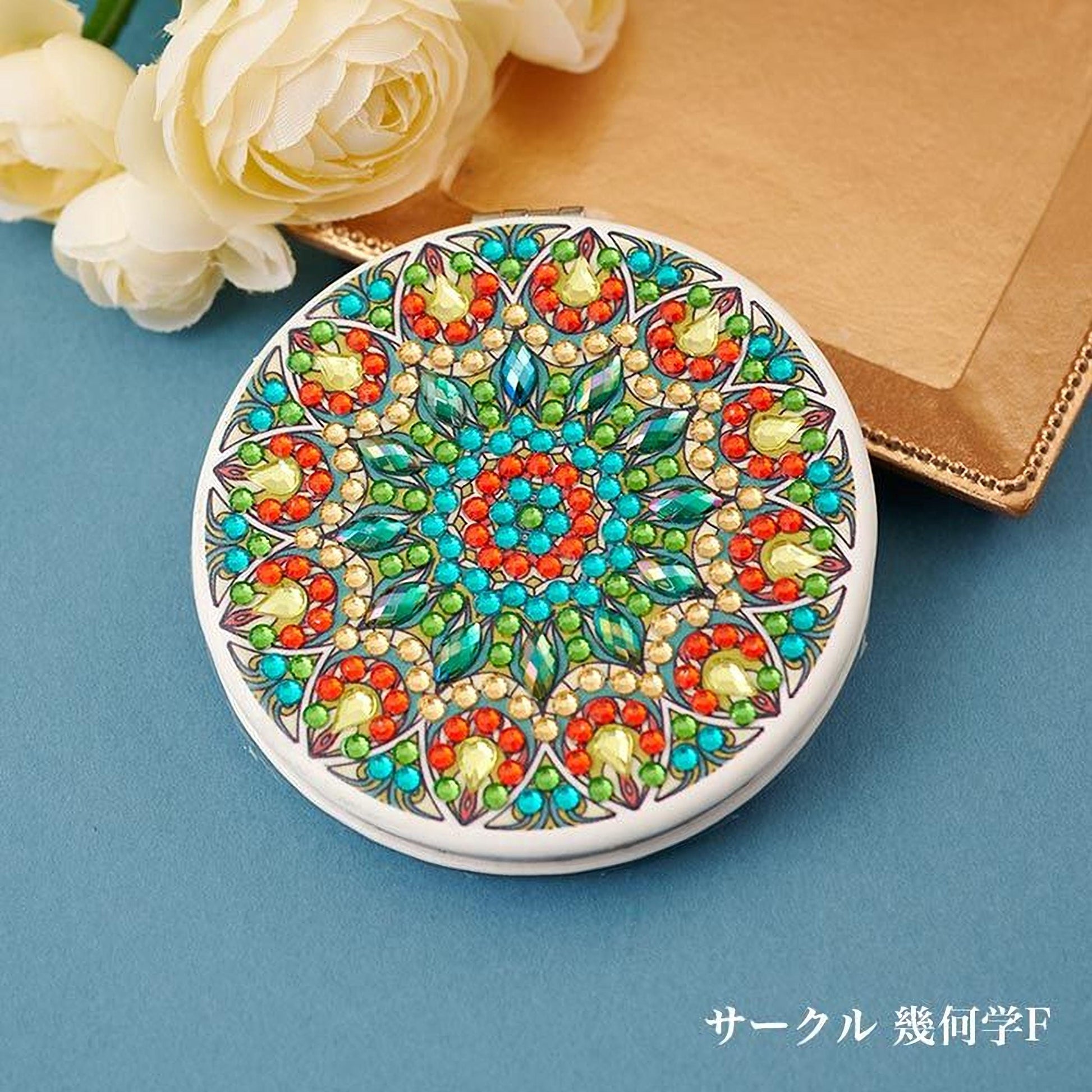 A decorative round mirror with a colorful, intricate design featuring various shapes, patterns, and gemstones. The mirror is placed on a blue surface, with a bouquet of white flowers in the background.