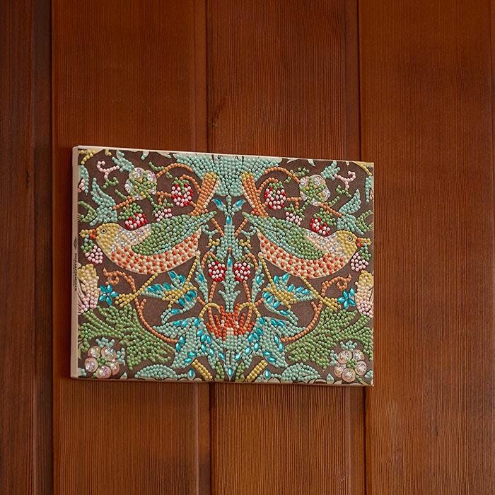 a vibrant, colorful, and intricate embroidered piece of art, which appears to be a decorative canvas or frame hanging on a wooden wall.
