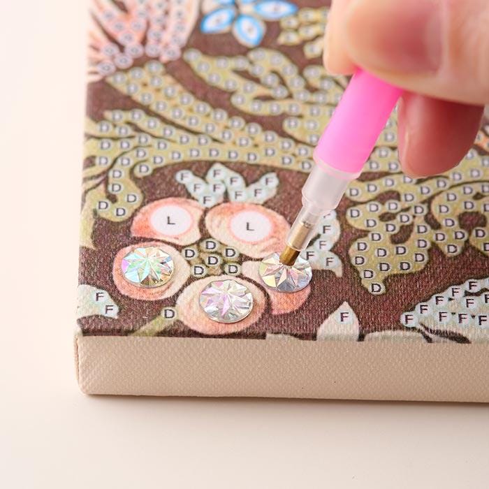 A hand is using a pink pen to work on a canvas with a floral pattern, adding rhinestones or other embellishments to it.