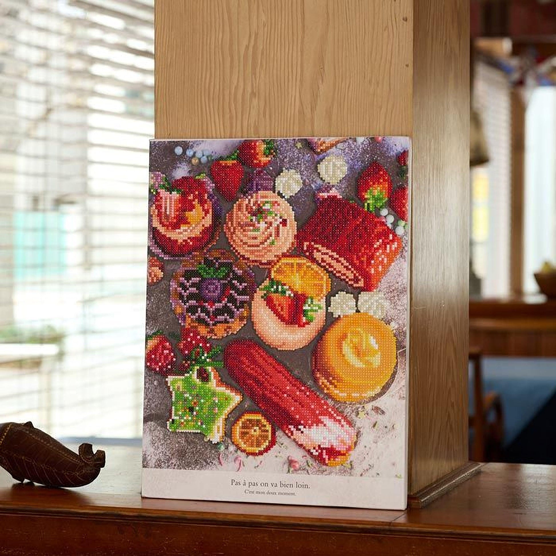 A colorful embroidered canvas depicting various desserts, including cakes, pastries, and fruits, is displayed on a wooden surface.