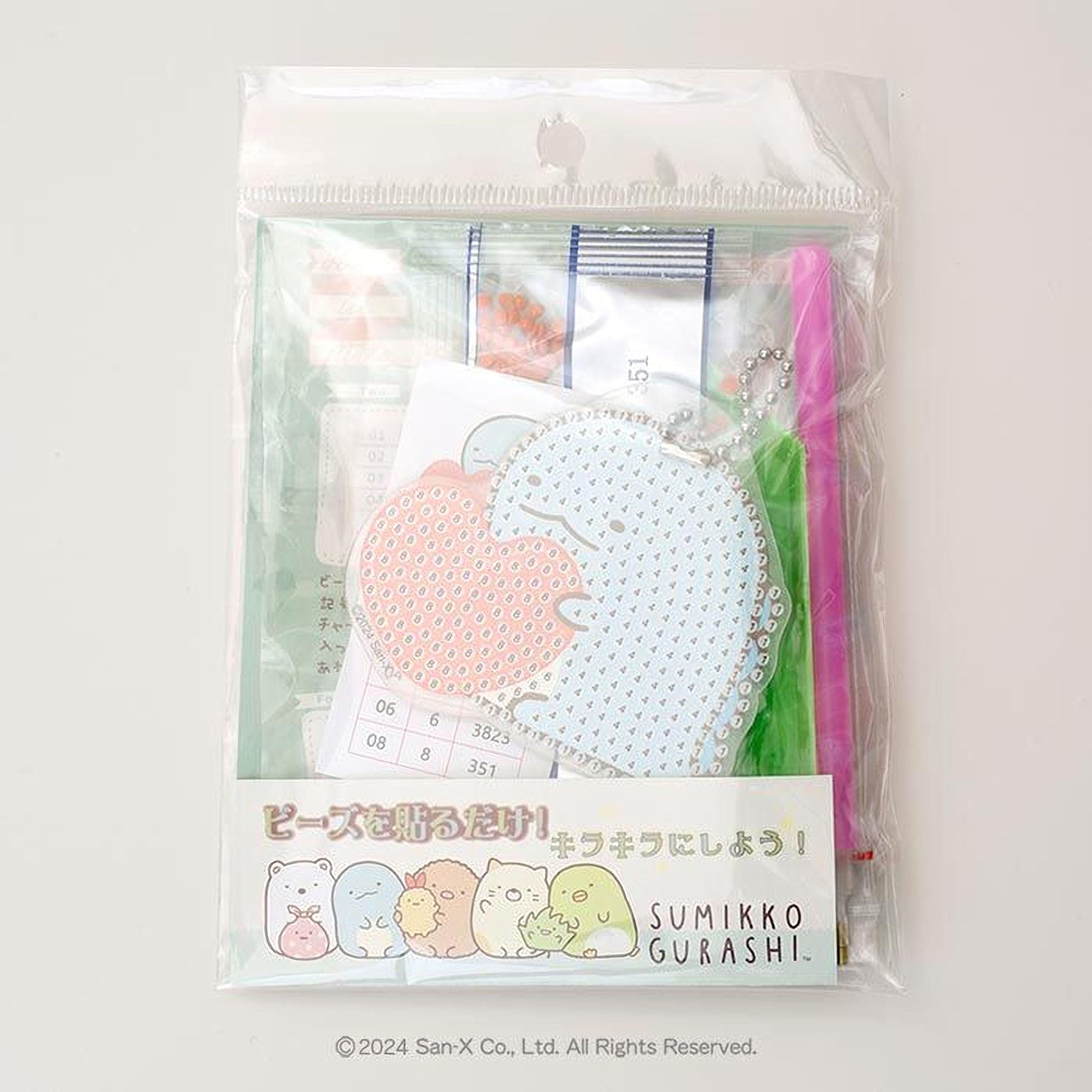a package with a clear plastic bag, which appears to be a gift set. The package contains a keychain, a pen, and a small toy. The keychain has a heart-shaped design, and the pen is pink. The toy is a small stuffed animal. The package also includes a label with Japanese text and a drawing of a group of cartoon characters.