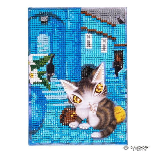 A diamond-studded embroidery depicting a cat with large, yellow eyes and a striped pattern, sitting on a blue surface with a house and other objects in the background.