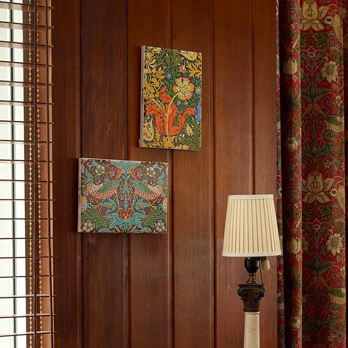 a wooden wall with two framed artworks hanging on it, one depicting a dragon and the other a floral design. In the foreground, there is a lamp with a white shade, and a window with wooden blinds can be seen on the left side of the image.