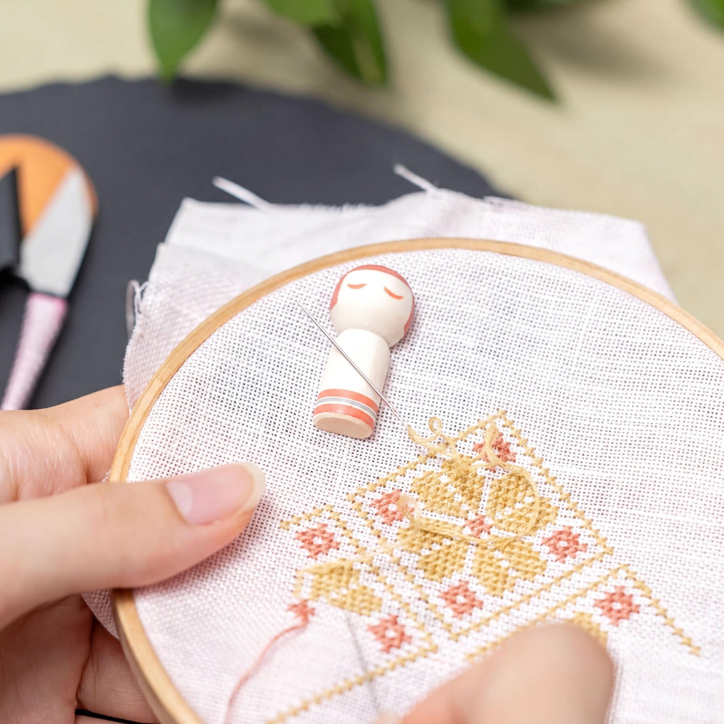 A person's hand holding a hoop with a small embroidery figure on it, surrounded by a cross-stitch design.