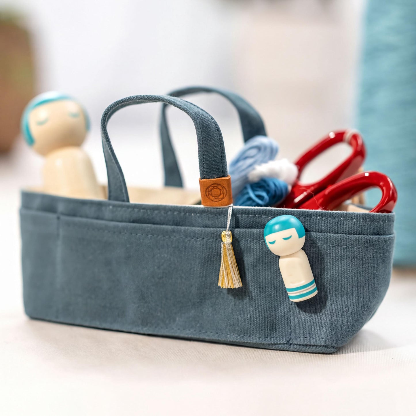 A small blue tote bag with a tassel hanging from the handle, containing various craft supplies such as scissors and a small wooden doll.