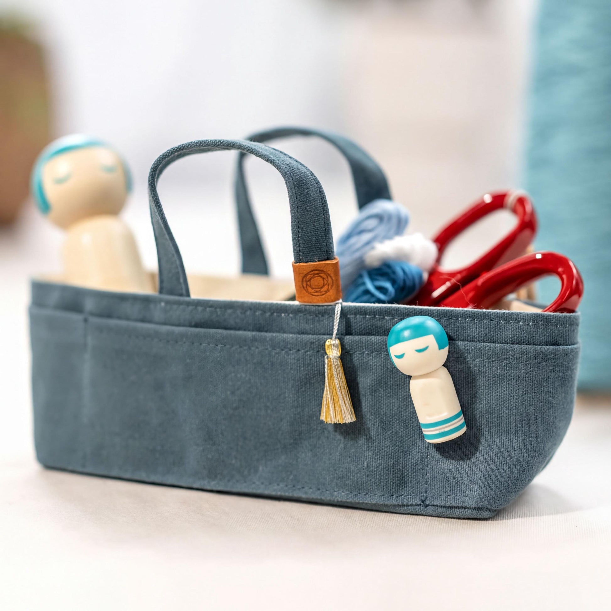 A small blue tote bag with a tassel hanging from the handle, containing various craft supplies such as scissors and a small wooden doll.
