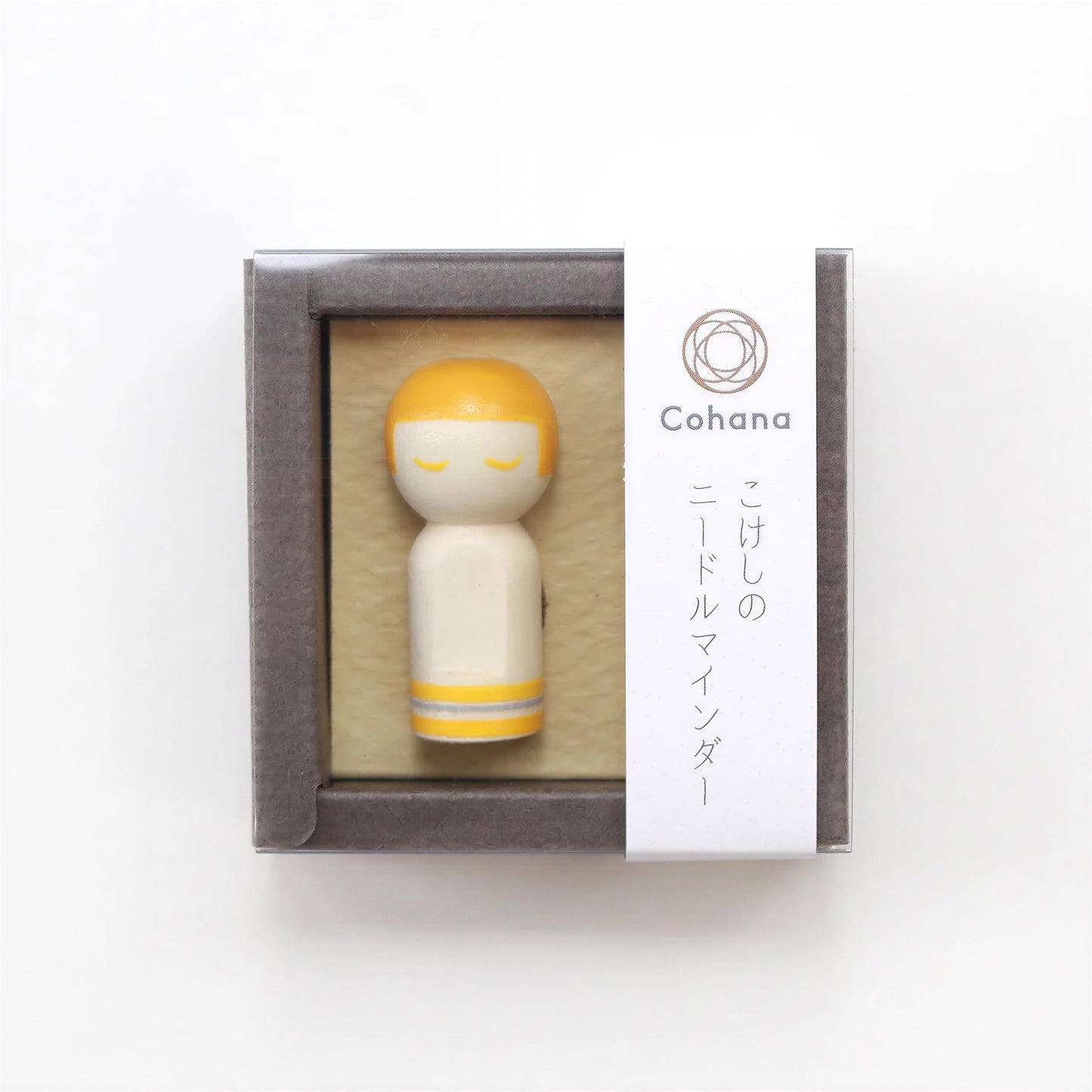A small wooden figurine with a yellow head and white body, displayed in a gray box with Japanese text and a circular logo.