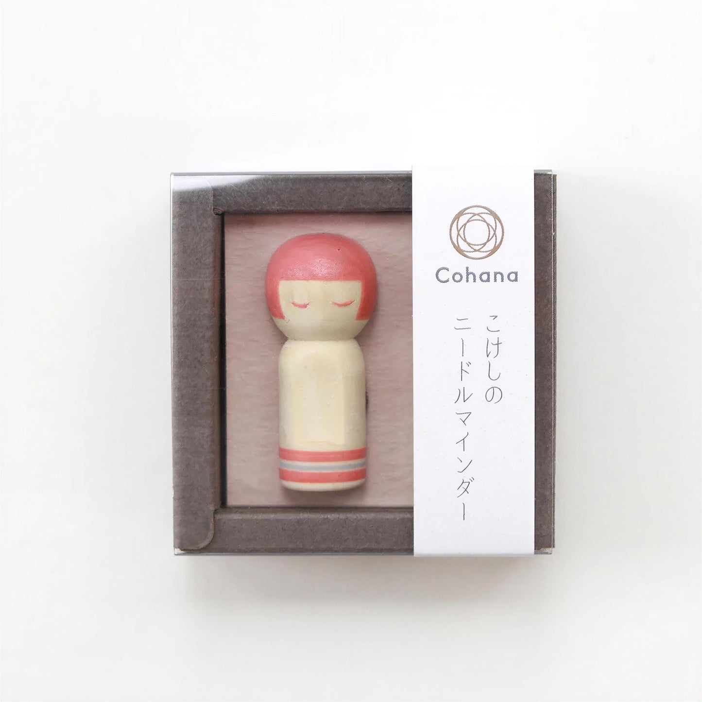 A small wooden doll with a pink head and red hair, displayed in a clear plastic box with Japanese text and a logo.