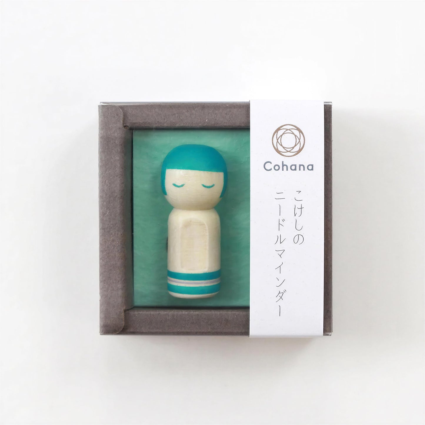 A small wooden doll with a teal head and white body, displayed in a gray box with a white label.