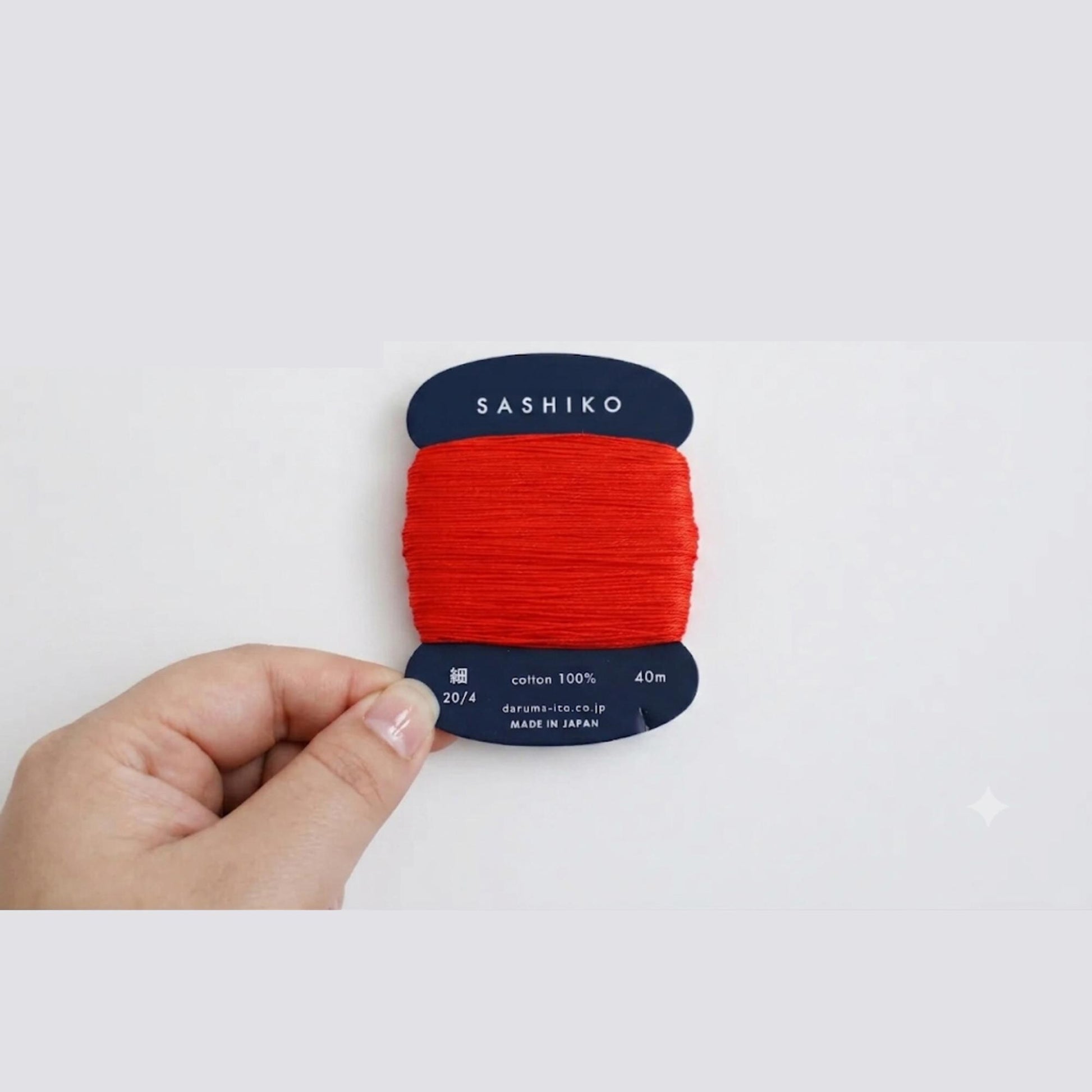 A hand is holding a spool of red thread with the brand name "SASHIKO" printed on it.