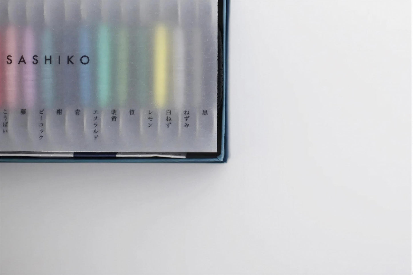 A transparent box containing a collection of colorful markers or pens, with the brand name "ASHIKO" printed on the side.