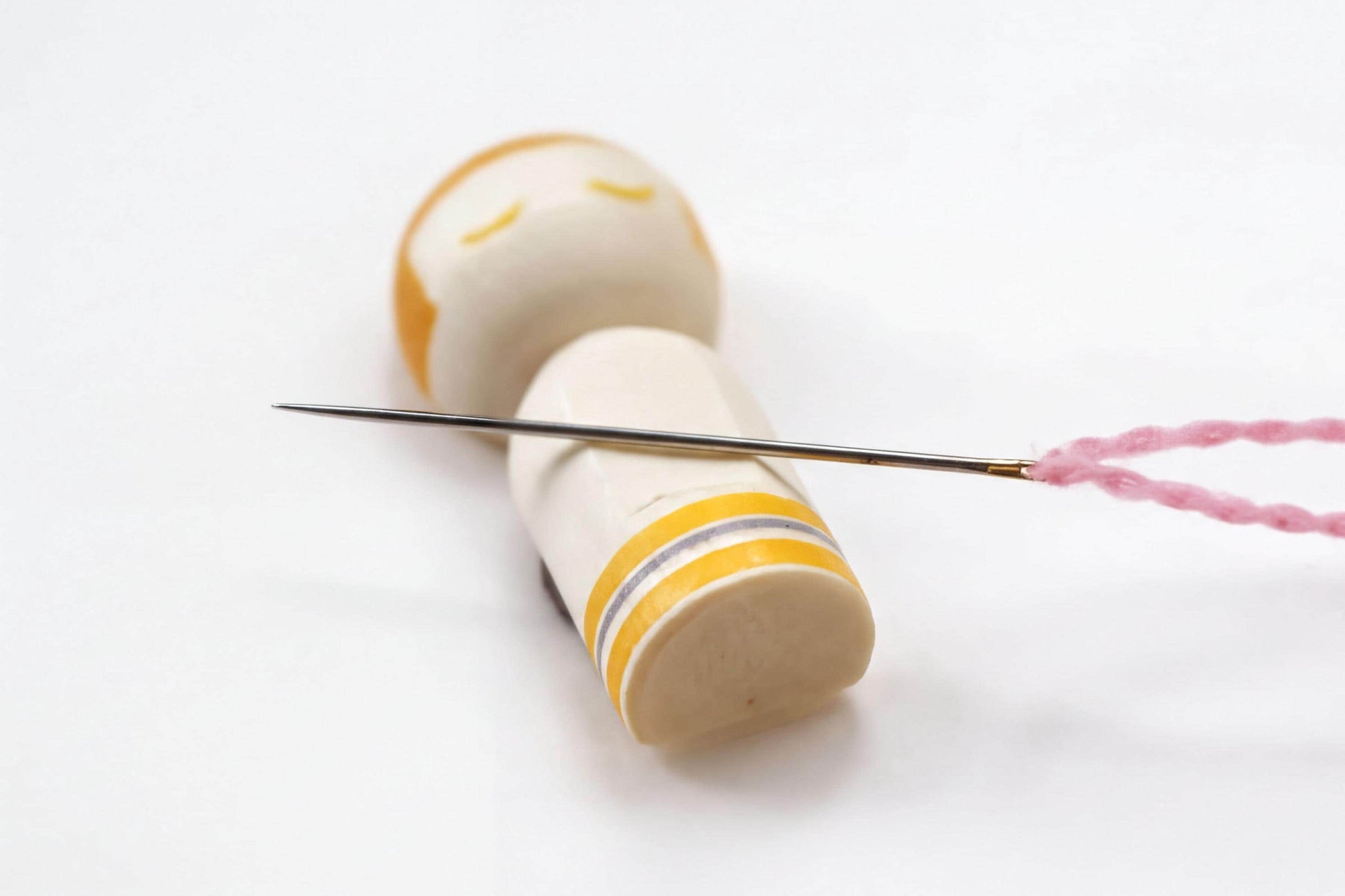 A wooden doll with a yellow and white striped band is lying on its back, with a needle and pink thread attached to it.