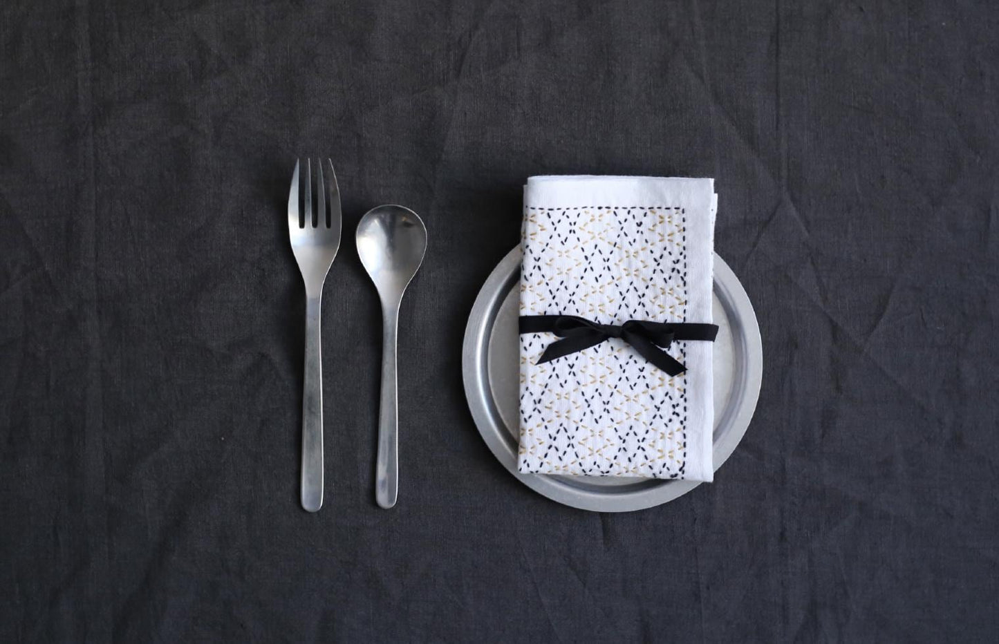A silver fork, spoon, and a napkin with a black bow tie are arranged on a dark grey tablecloth.