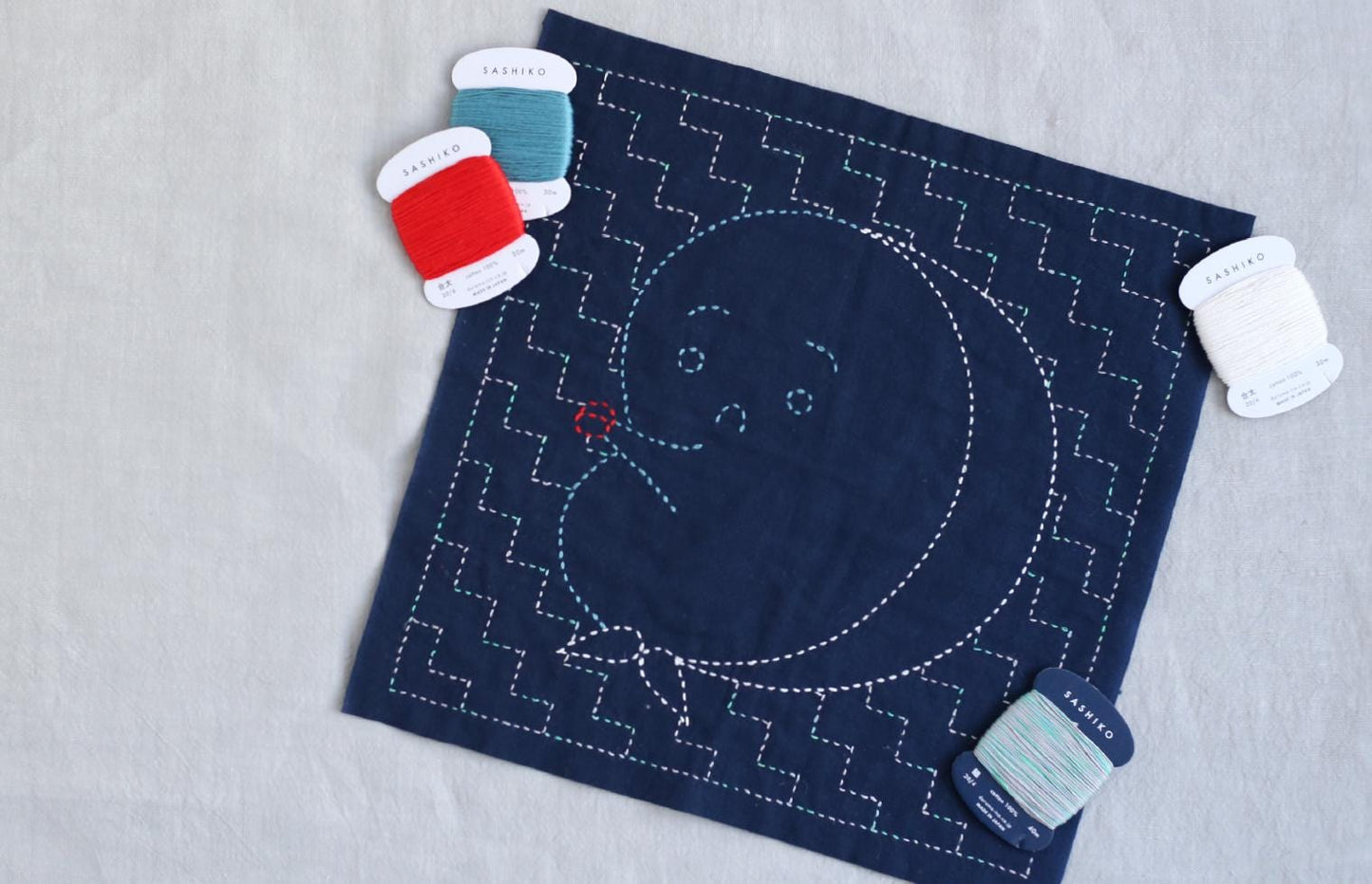 a blue square with a white and red thread design, along with some thread spools and a needle, suggesting a sewing or crafting project.