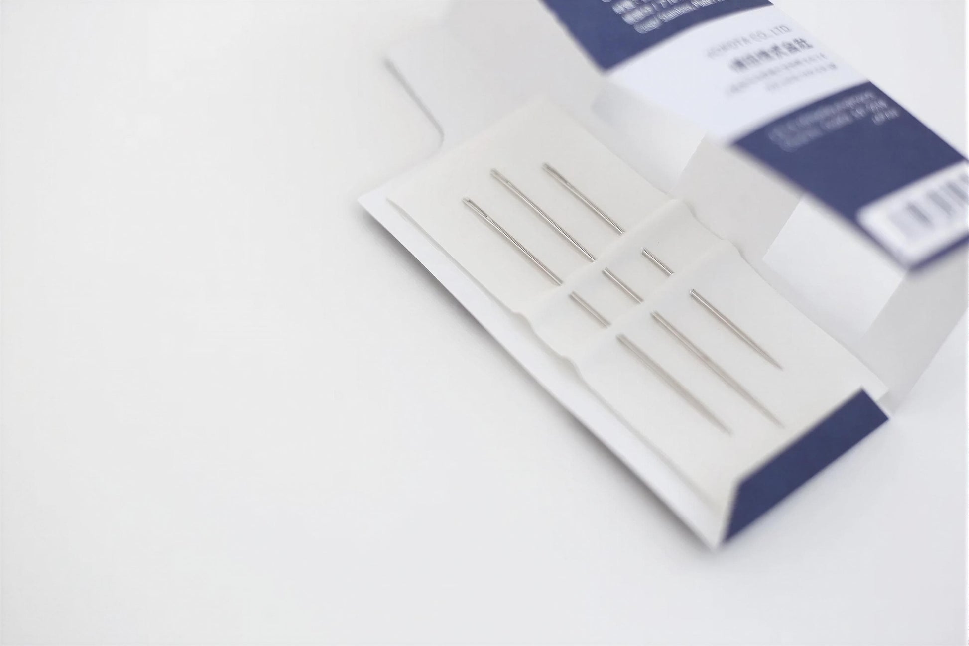 A close-up view of a white box containing a set of sewing needles, with the needles clearly visible and the box's label facing upwards.