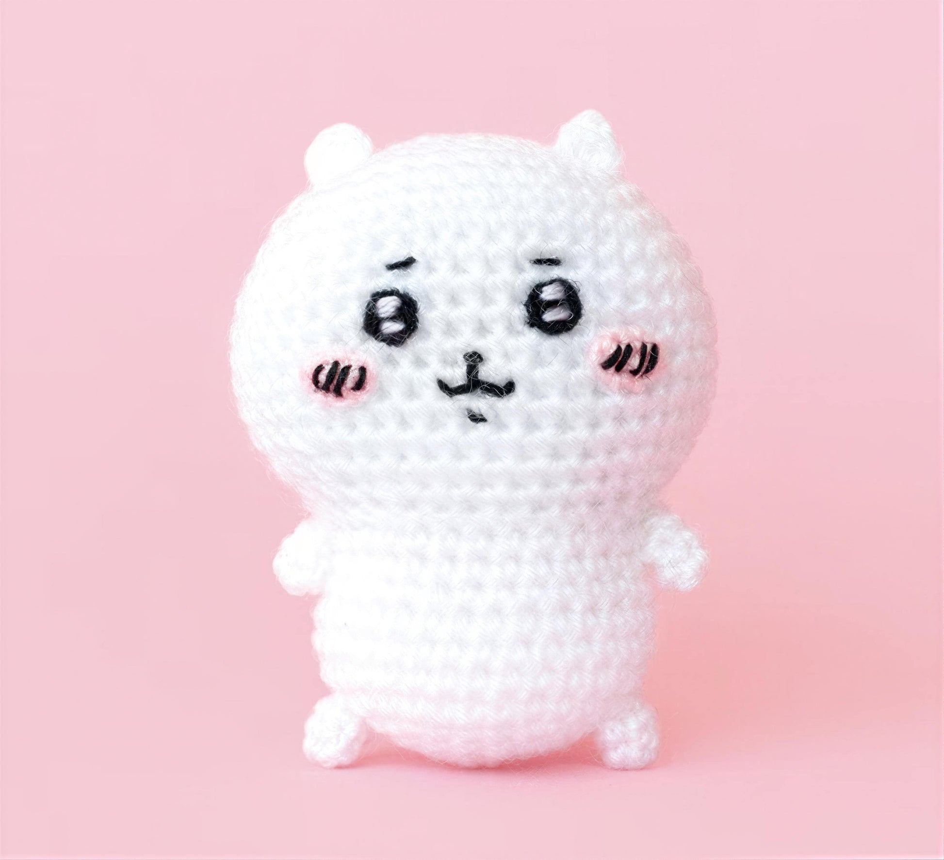 A white crocheted doll with large eyes and a small nose, standing on a pink background.