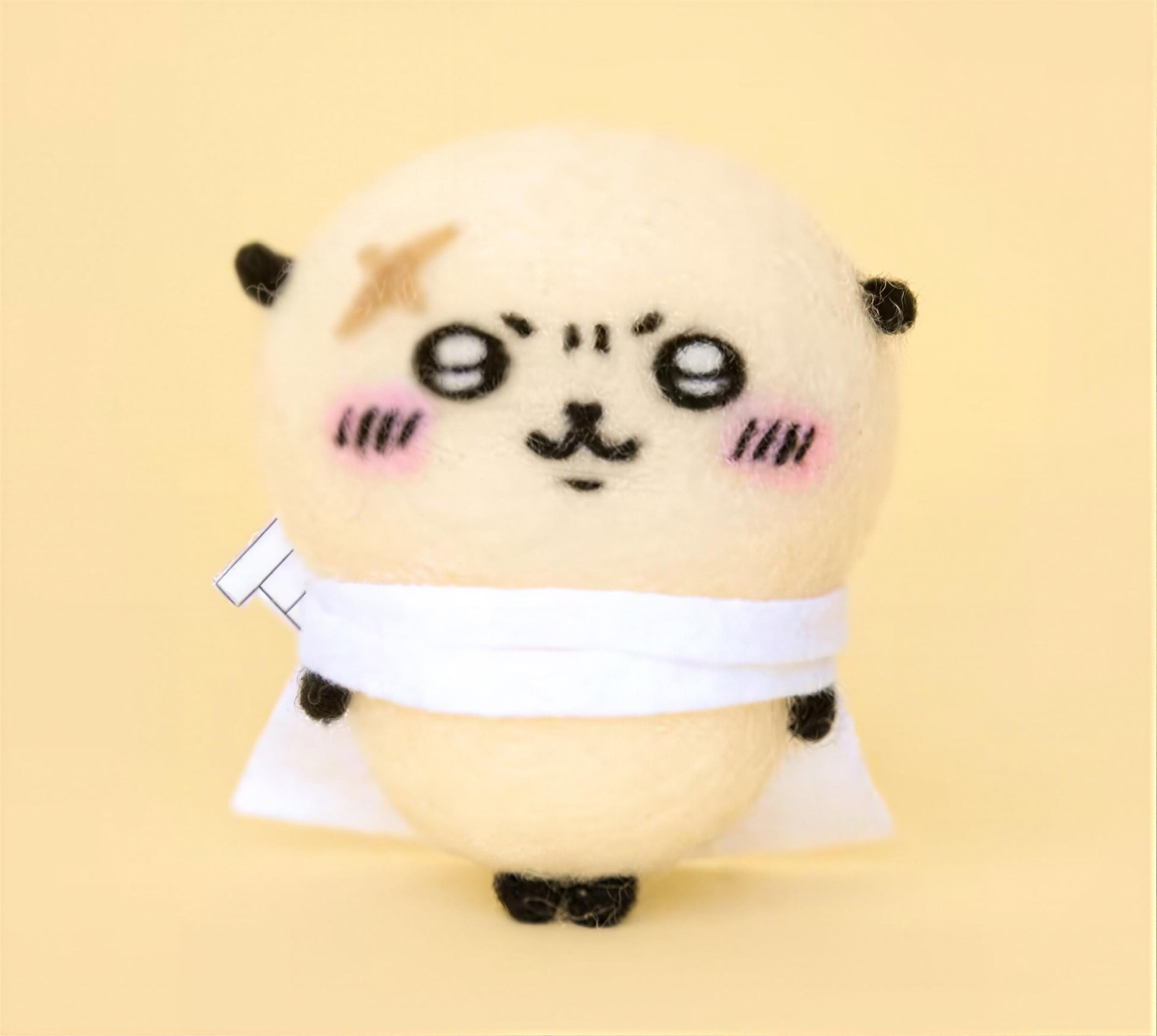 A cute, hand-made plush toy with a round body, large eyes, and a small nose, wearing a white scarf and standing on a yellow background.