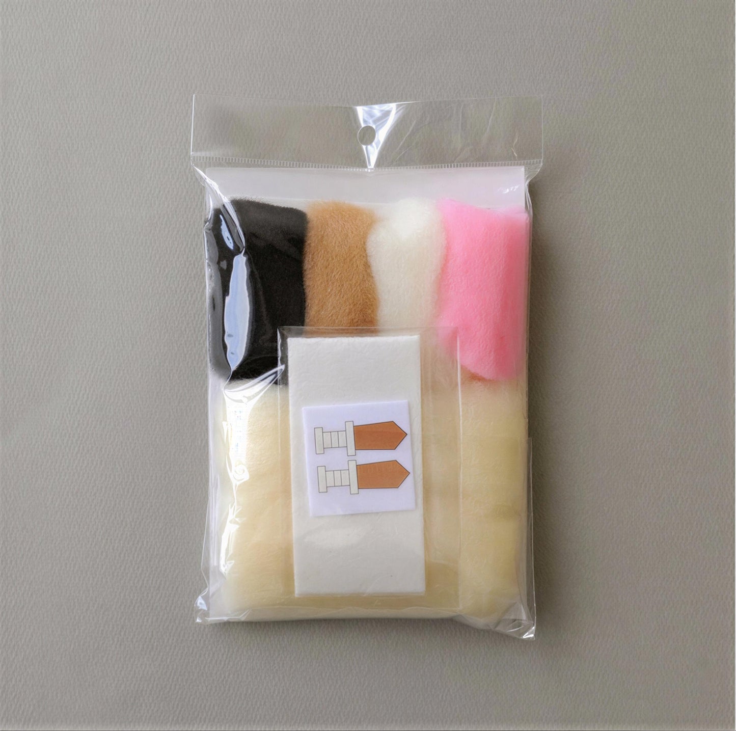 A clear plastic bag containing various colored felt pieces, including black, pink, and white, along with a label with a pair of scissors.