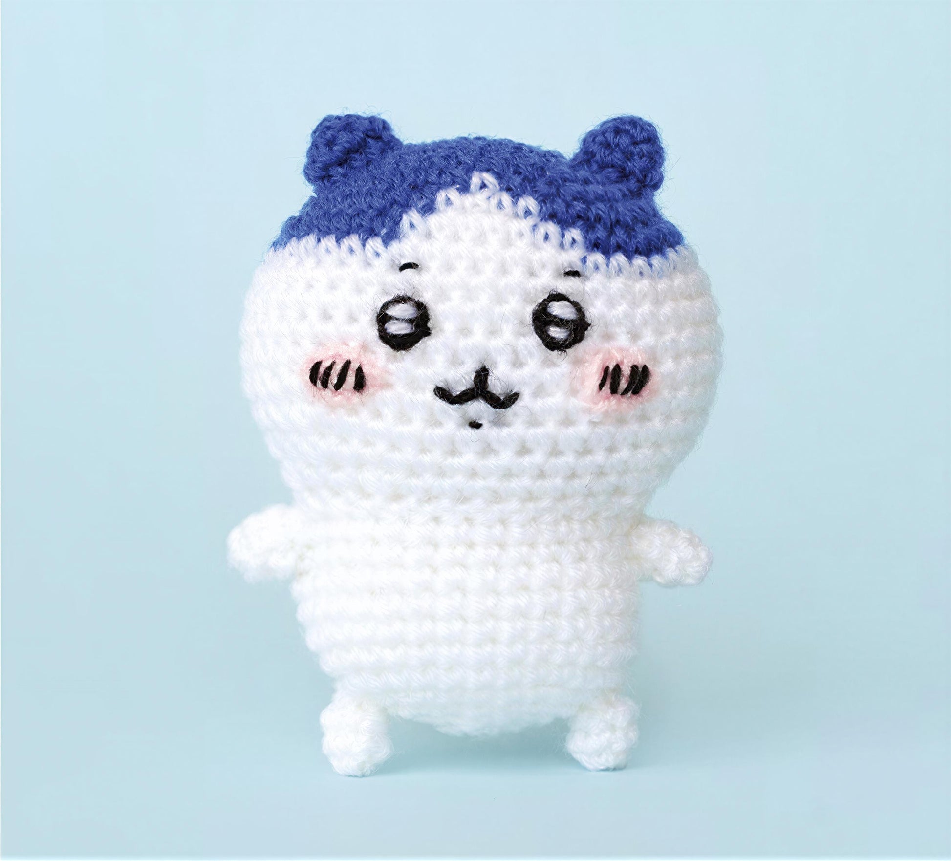 A crocheted white doll with blue ears and a black nose, standing on two legs against a light blue background.