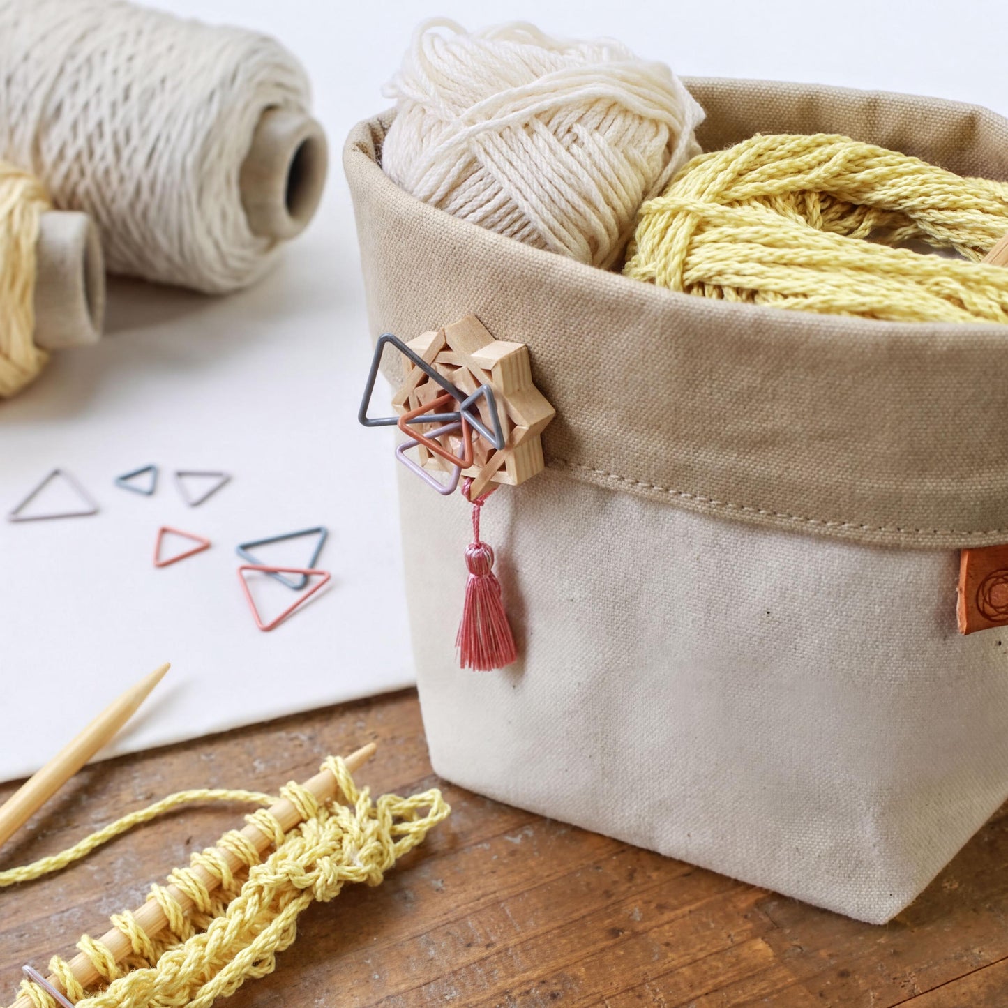 A beige basket filled with yarn and knitting needles sits on a wooden surface, with additional crafting supplies visible in the background.