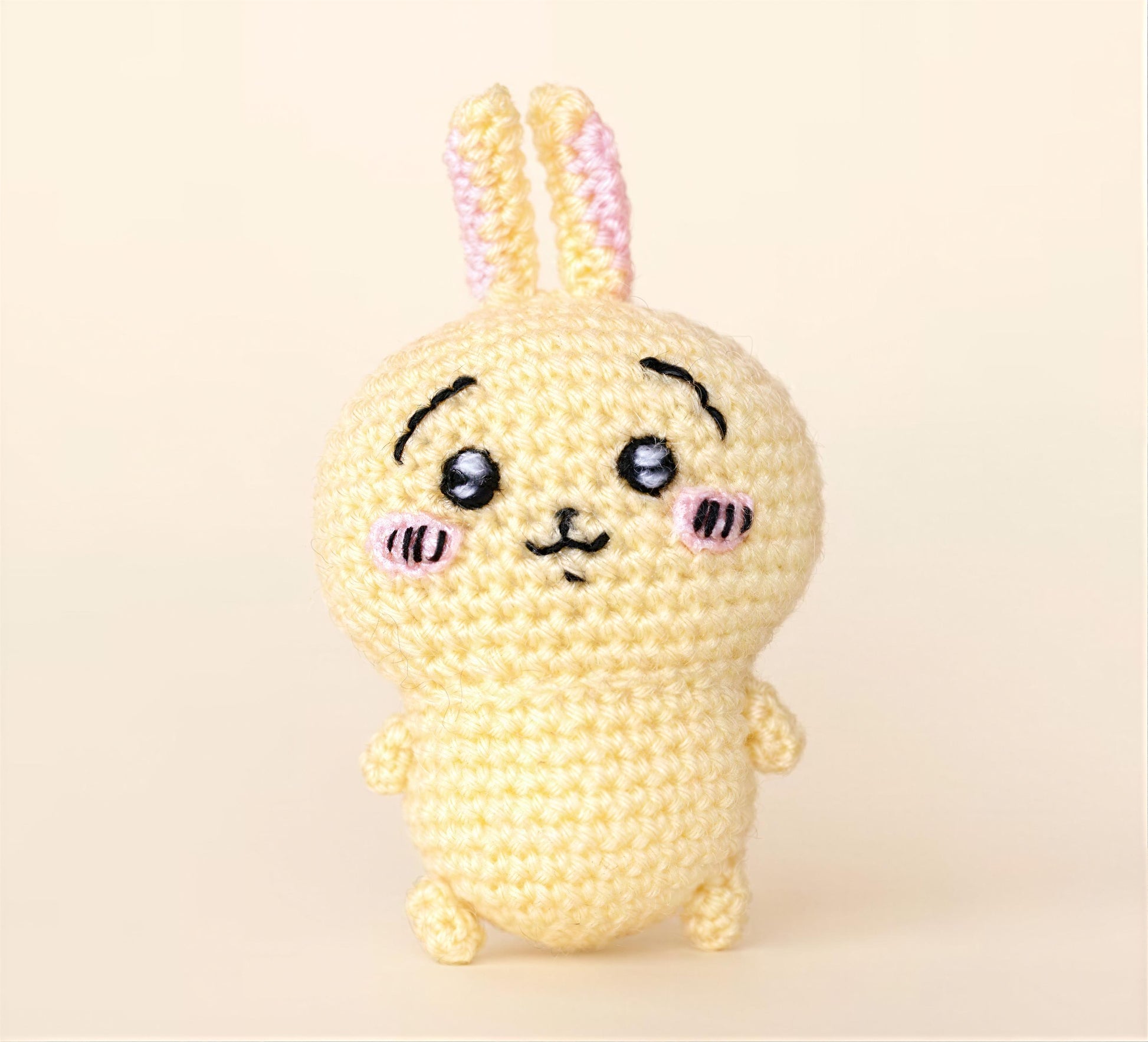 A crocheted toy rabbit with a cute face and pink ears is sitting on a plain background.