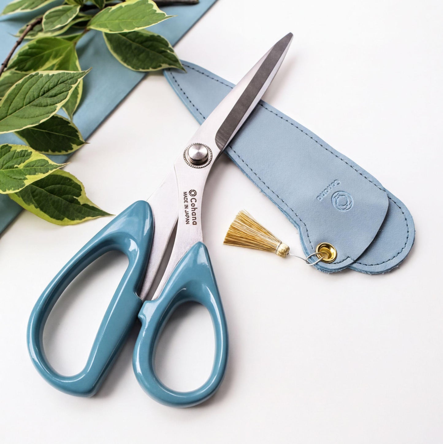 A pair of blue-handled scissors with a tassel on the side, placed on a white surface next to a small blue leather case.