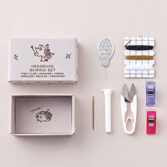 A sewing kit containing various tools and materials, including a box, scissors, pins, thread, needles, and a thimble, arranged on a light background.