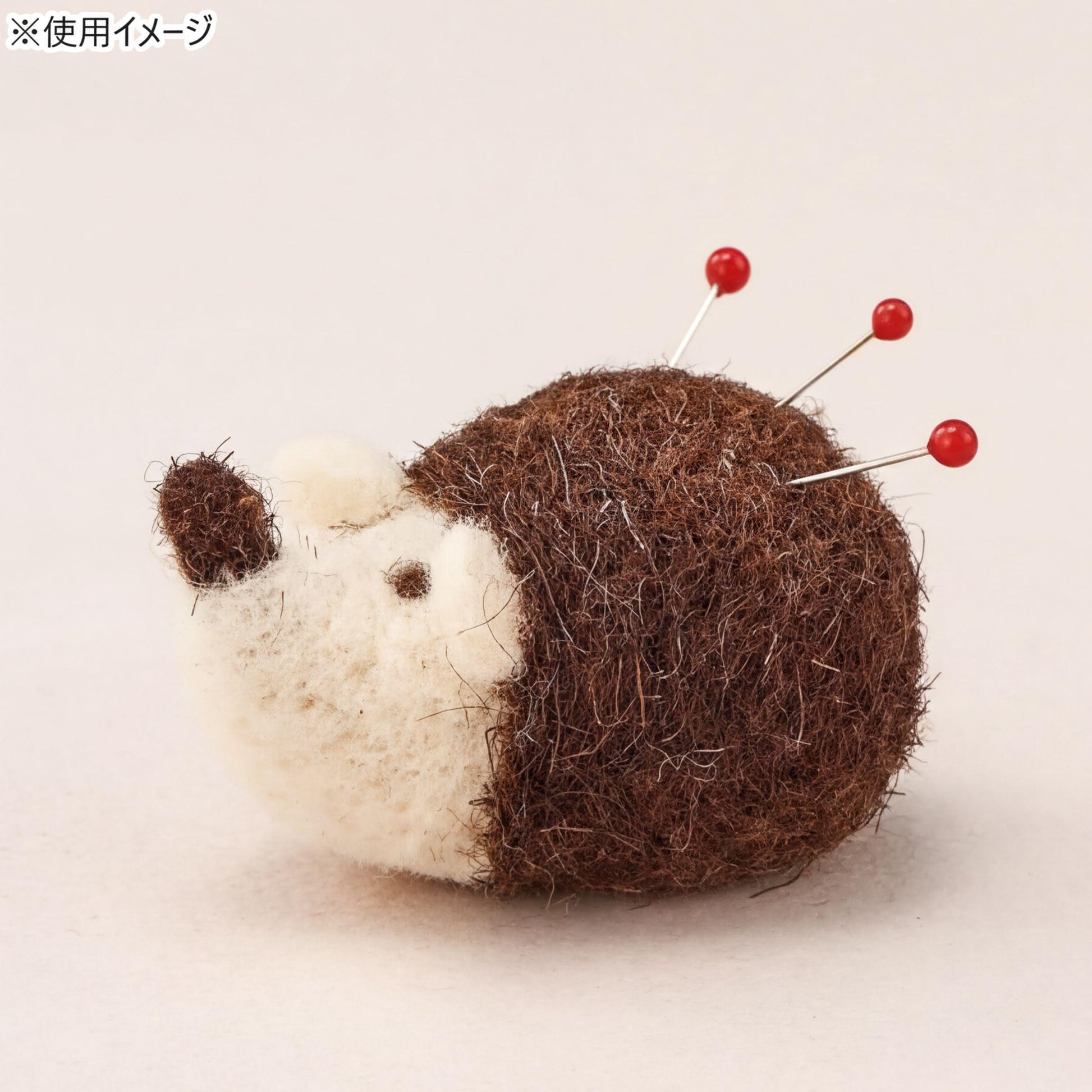 A brown and white felt hedgehog pin cushion with red pins protruding from its head.