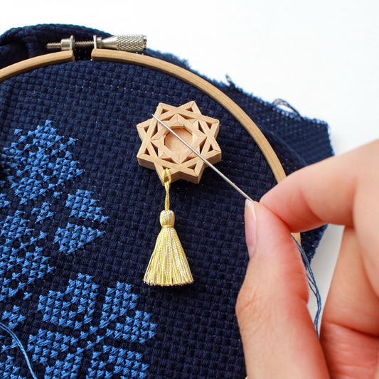 A hand is holding a needle and thread, stitching a wooden star-shaped object onto a blue fabric with a cross-stitch pattern.