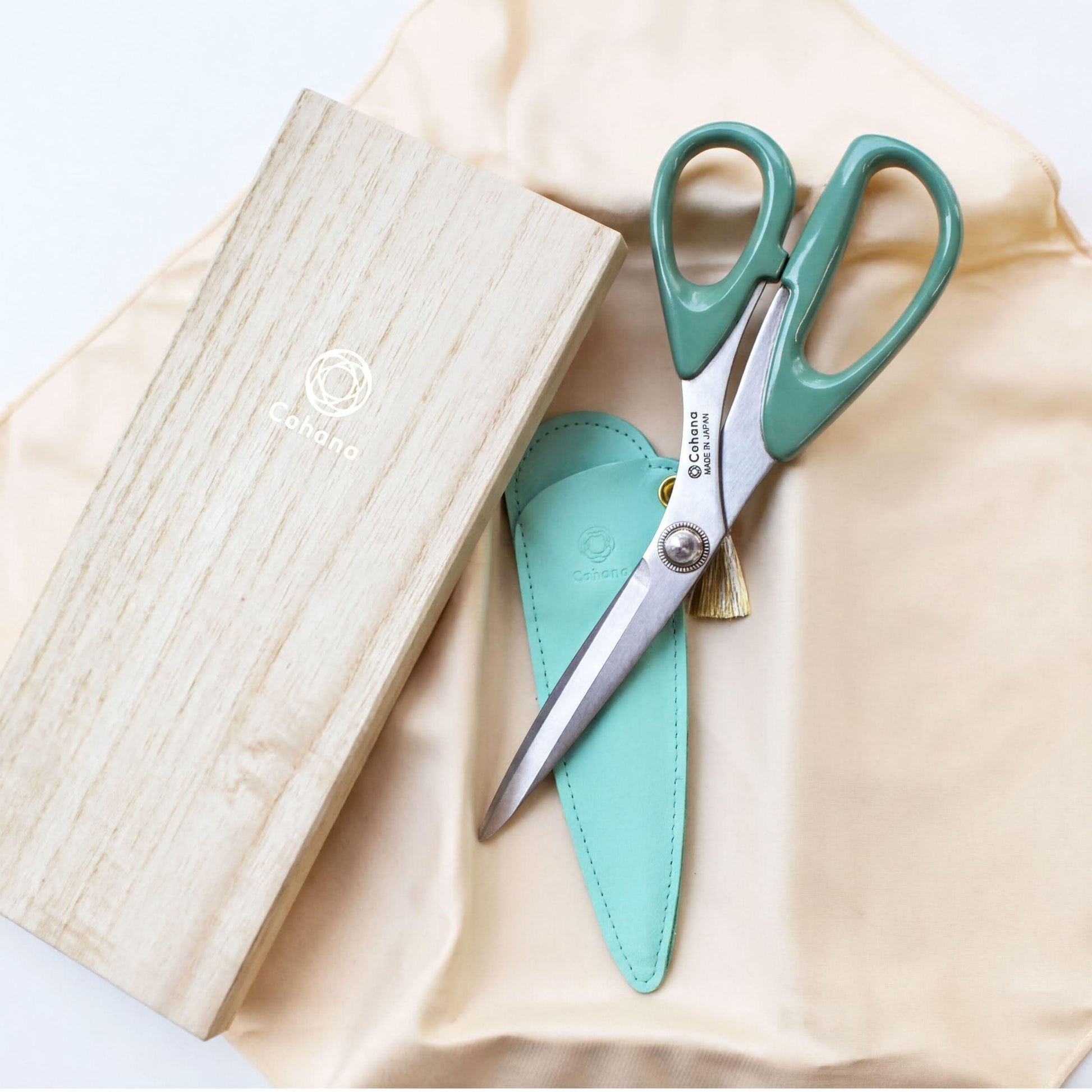 A pair of green scissors with a wooden box and a teal-colored pouch are placed on a beige fabric.