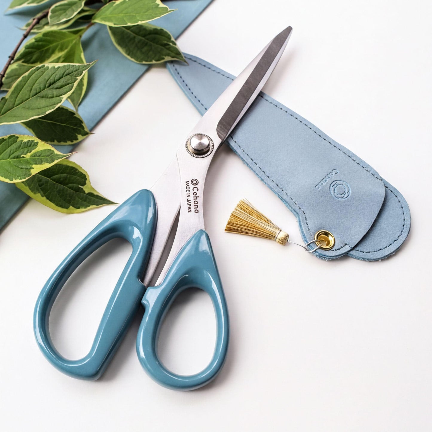 A pair of blue-handled scissors with a tassel on the handle, placed on a white surface next to a small blue leather case.