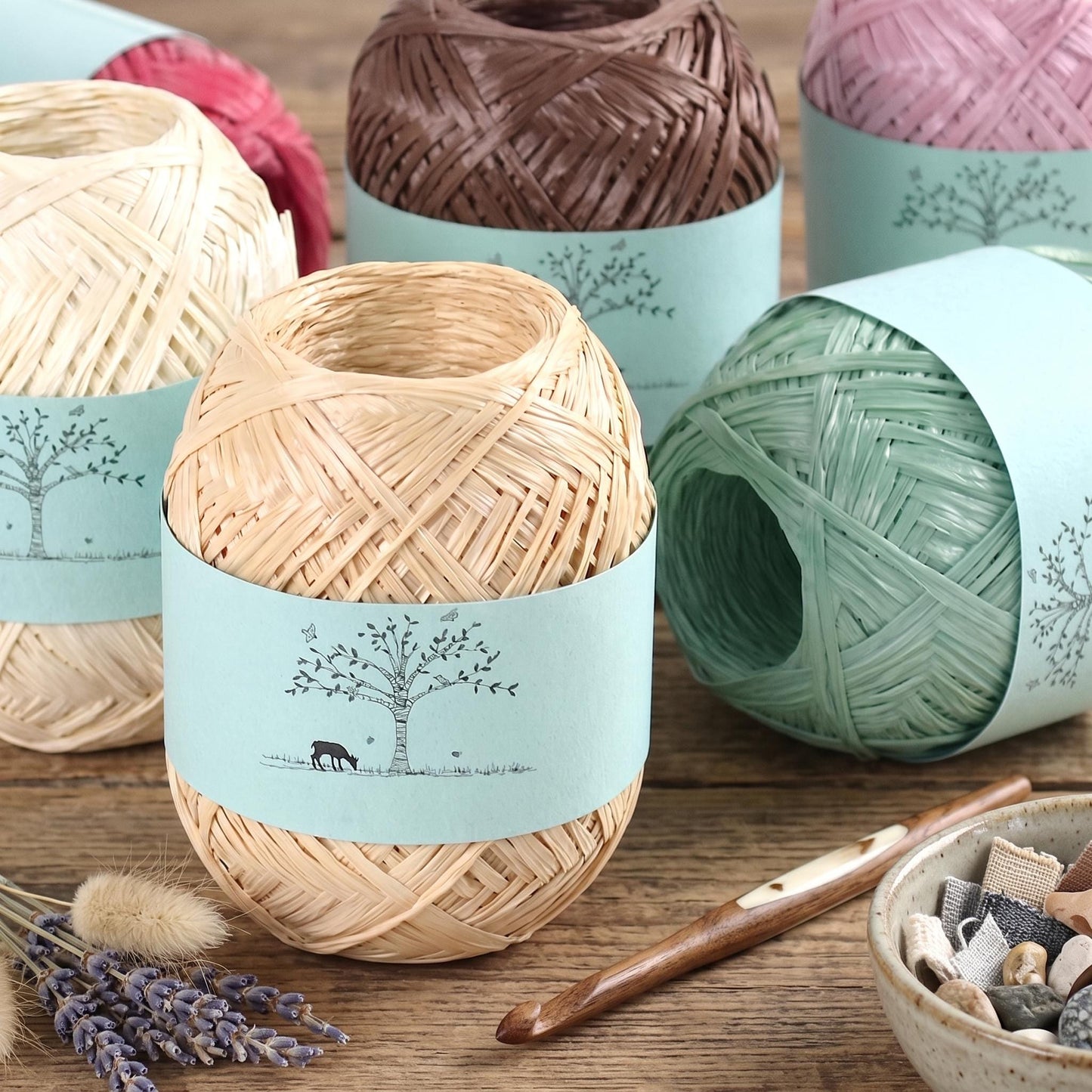 a collection of yarn balls wrapped in blue and green wrapping paper, along with crochet hooks and a bowl of small rocks, all arranged on a wooden surface.
