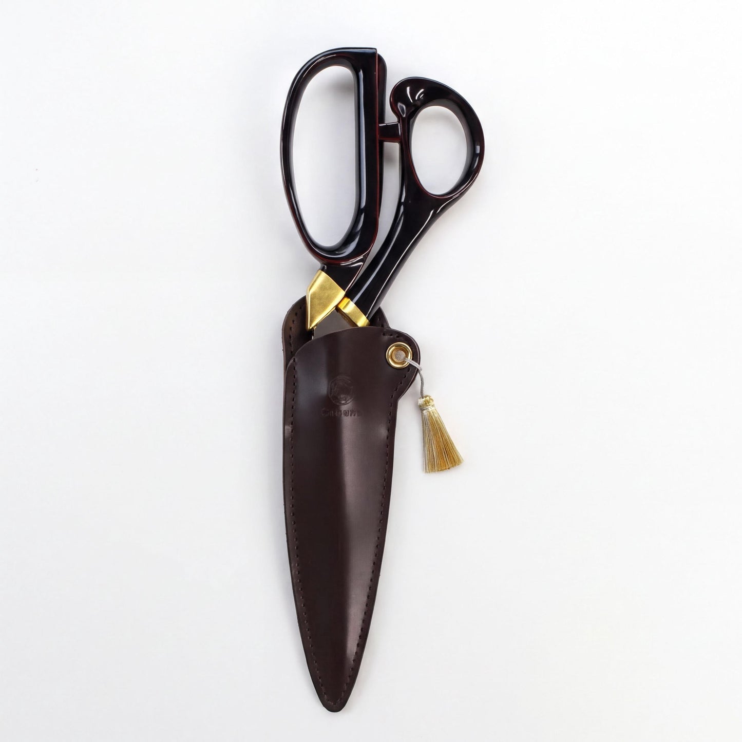 A pair of black scissors with gold accents, including a tassel, is displayed in a brown leather pouch against a white background.