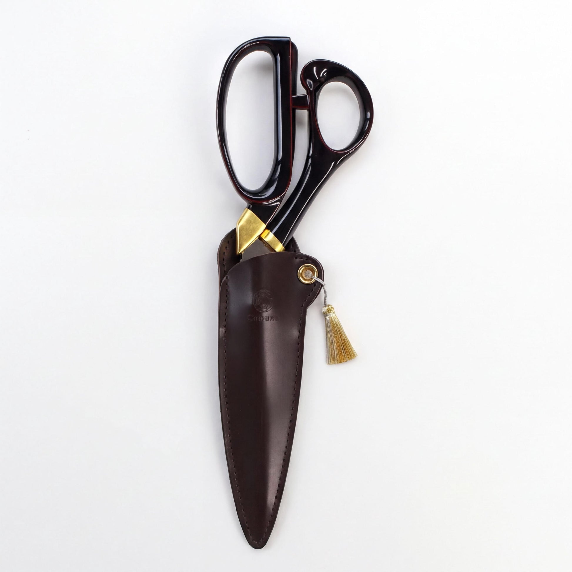 A pair of black scissors with gold accents, including a tassel, is displayed in a brown leather pouch against a white background.