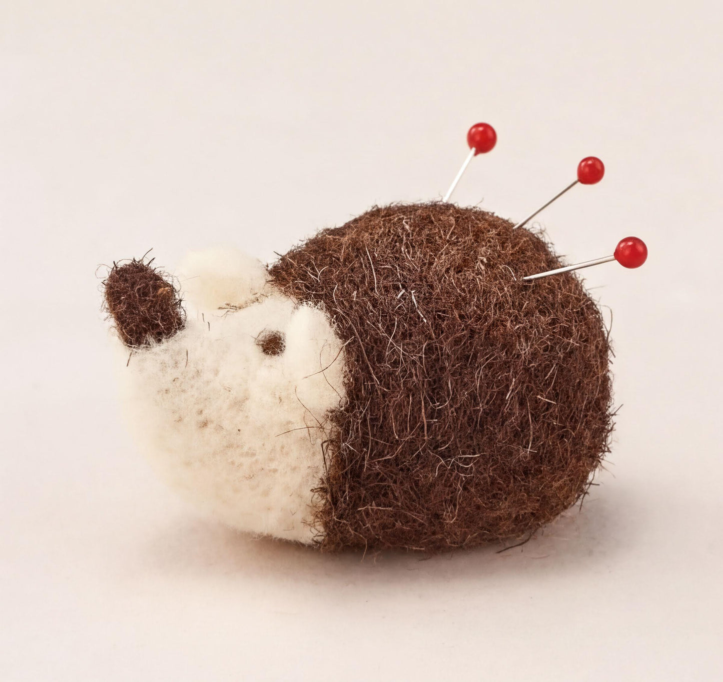 A brown and white felt hedgehog pin cushion with red pins protruding from its head.