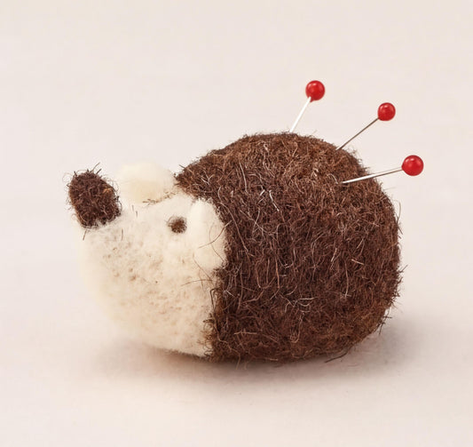 A brown and white felt hedgehog pin cushion with red pins protruding from its head.