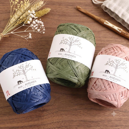 Three spools of yarn with labels for eco-friendly materials are arranged on a wooden surface, accompanied by a crochet hook and some flowers.