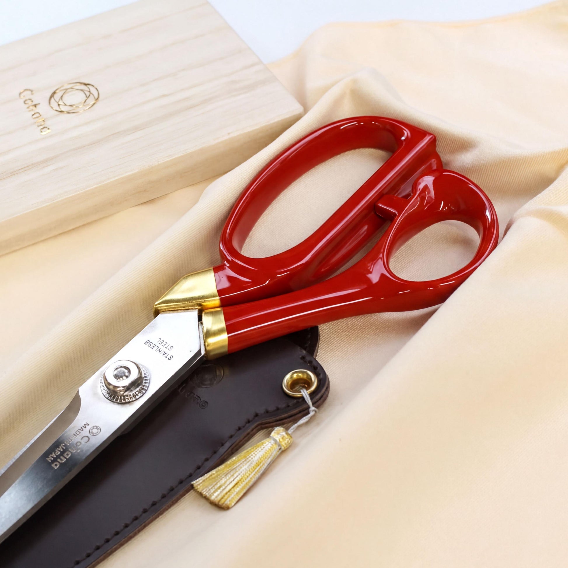 A pair of red and gold scissors with a black leather case and a gold tassel is shown on a beige fabric background.