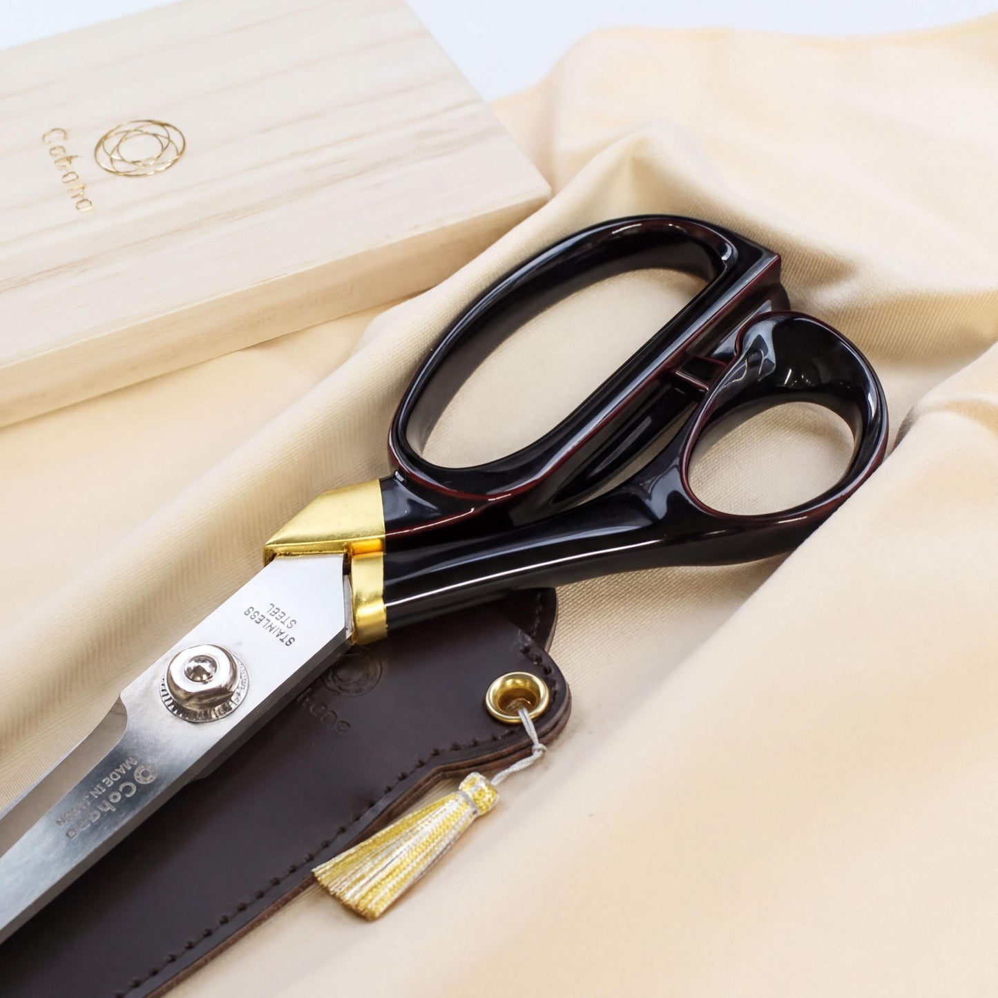 A pair of black and gold scissors with a wooden box and a tassel are displayed on a beige fabric background.