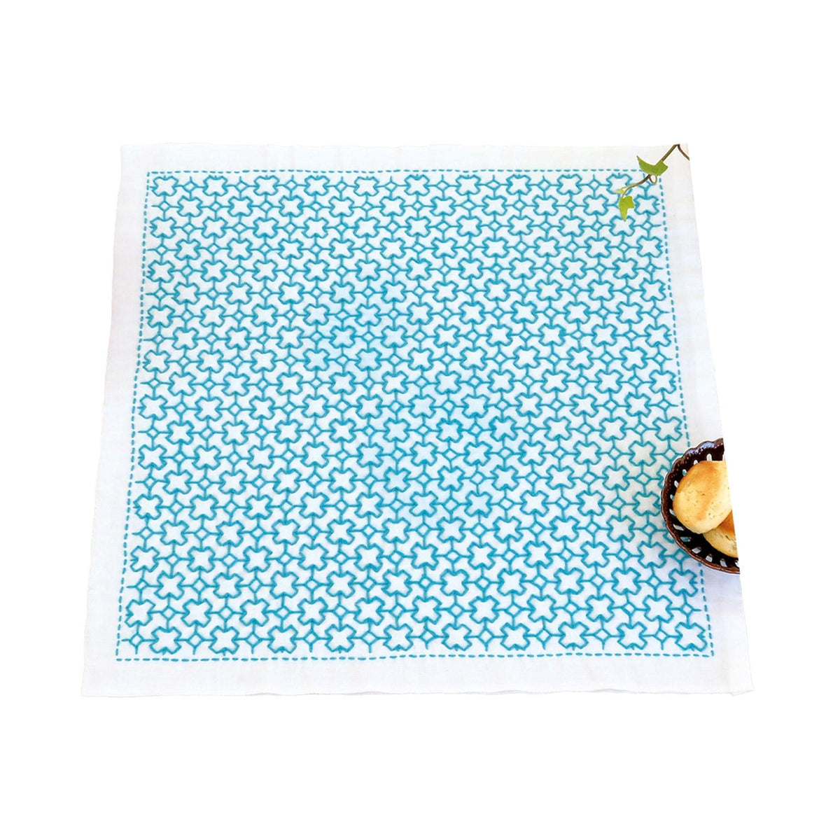 a square napkin with a blue and white geometric pattern, placed on a white surface next to a small bowl containing a piece of bread.
