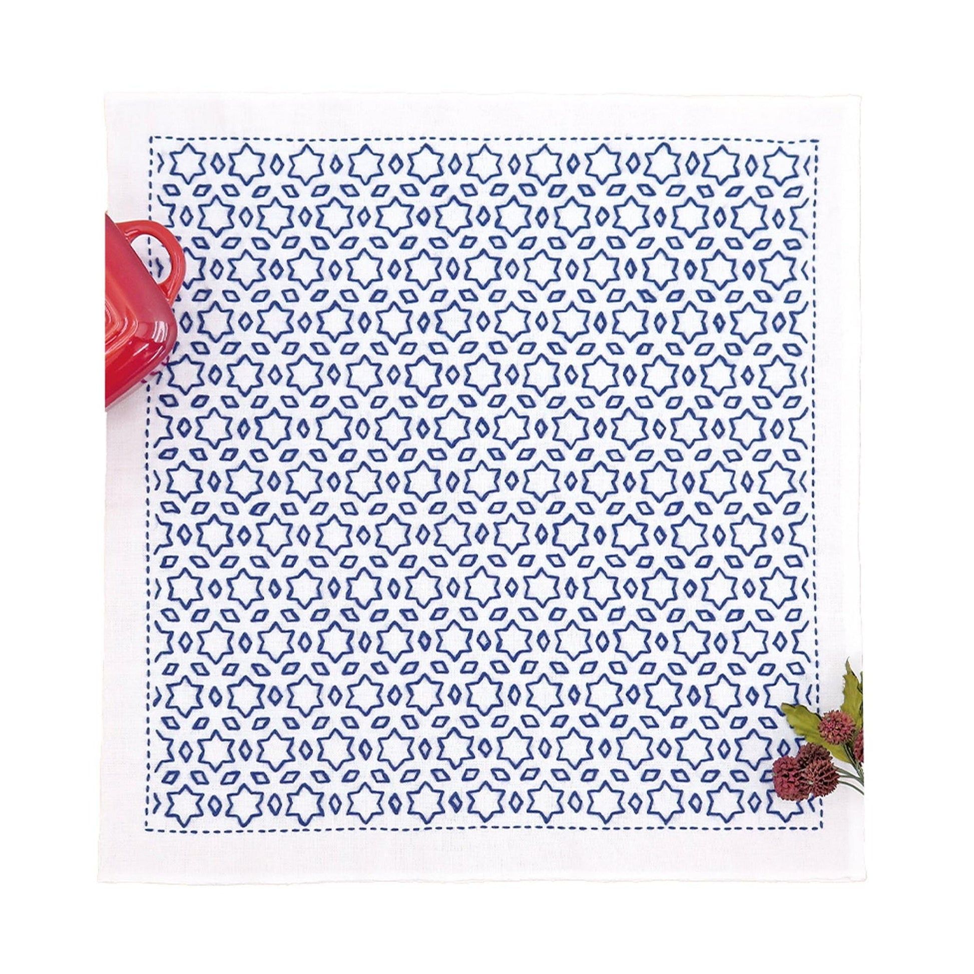 a square napkin with a blue and white star-patterned design, placed on a white surface. Next to the napkin, there is a red mug and a small plant.