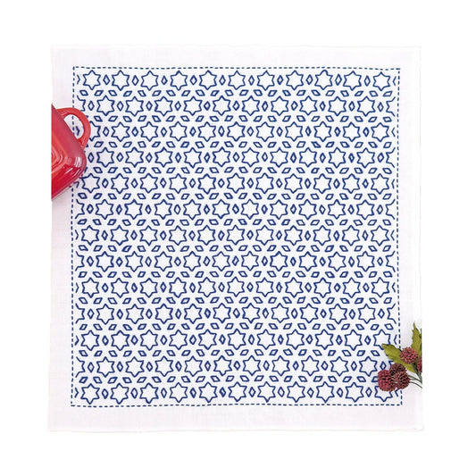 a square napkin with a blue and white star-patterned design, placed on a white surface. Next to the napkin, there is a red mug and a small plant.