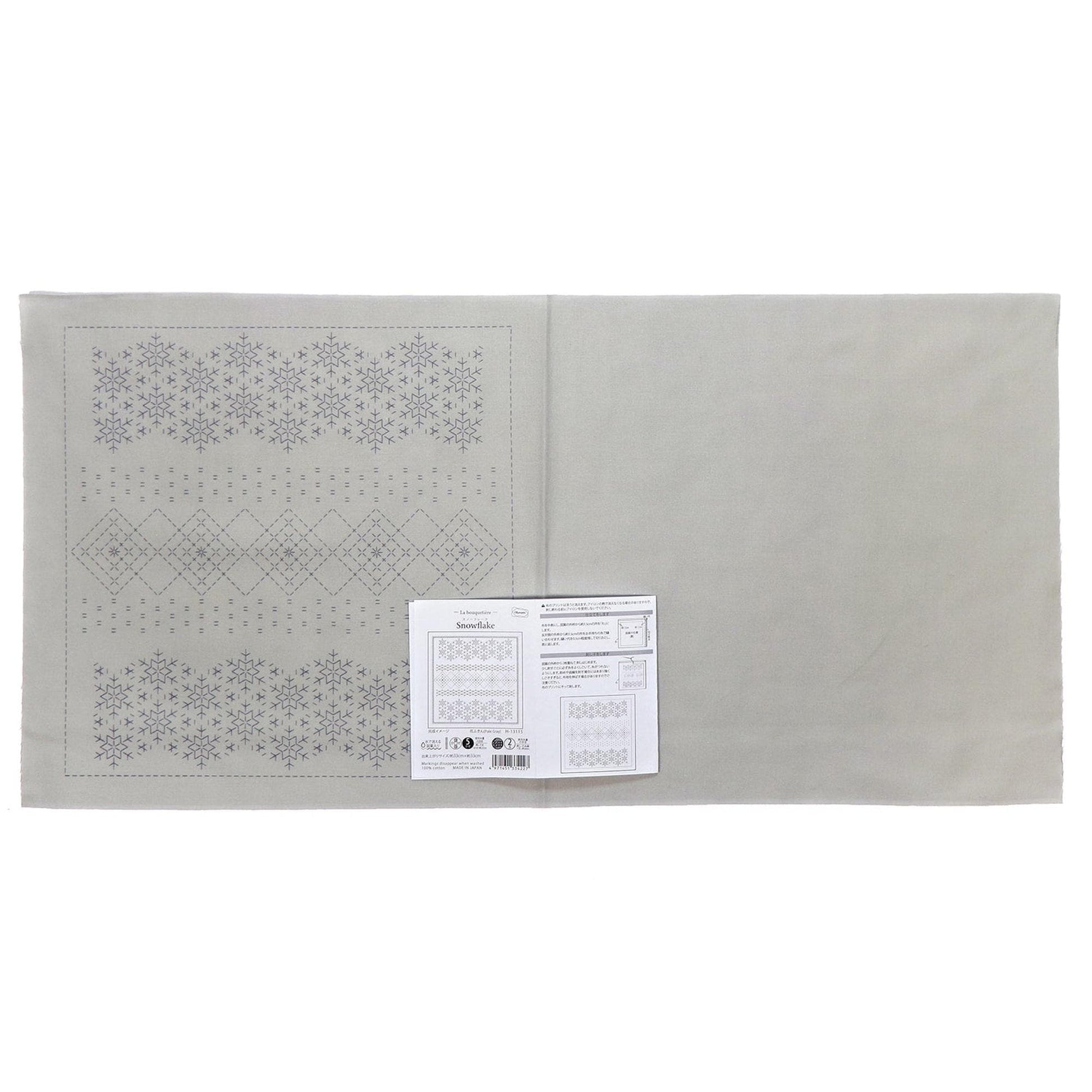 The image is a gray fabric with a repeating pattern of snowflakes on it. There is a white paper with text and a barcode attached to the fabric.