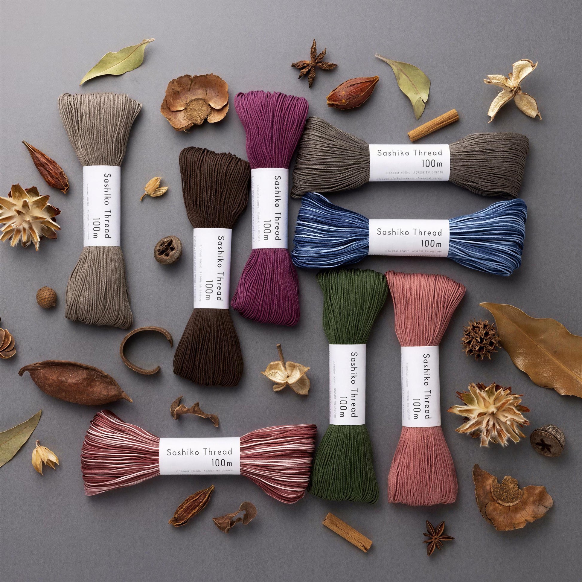 a collection of colorful thread spools, each labeled with the name of the thread type, arranged on a gray background alongside various natural elements such as dried leaves, pine cones, and star anise.