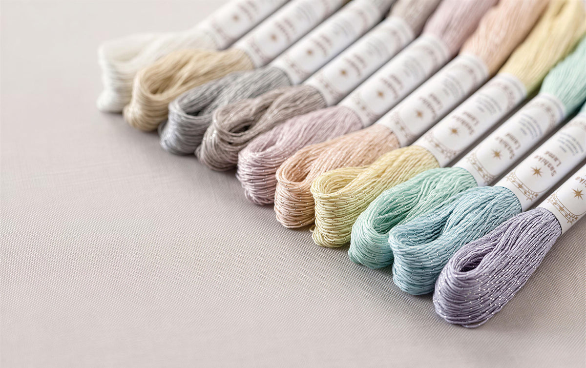 A collection of embroidery floss in various pastel colors, including white, light pink, light blue, light green, and light purple, arranged in a row on a light-colored surface.