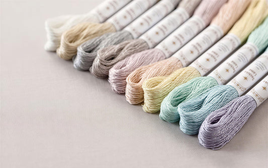 A collection of embroidery floss in various pastel colors, including white, light pink, light blue, light green, and light purple, arranged in a row on a light-colored surface.