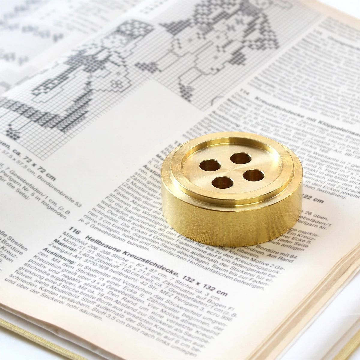 a close-up of a book with a cross-stitch pattern on the page, and a brass button placed on top of the page.