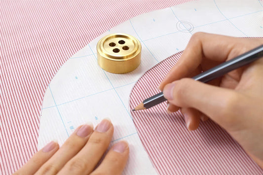 A person's hand is using a pencil to draw on a pattern on a red and white striped fabric, with a gold button and a piece of paper with a grid pattern visible in the background.