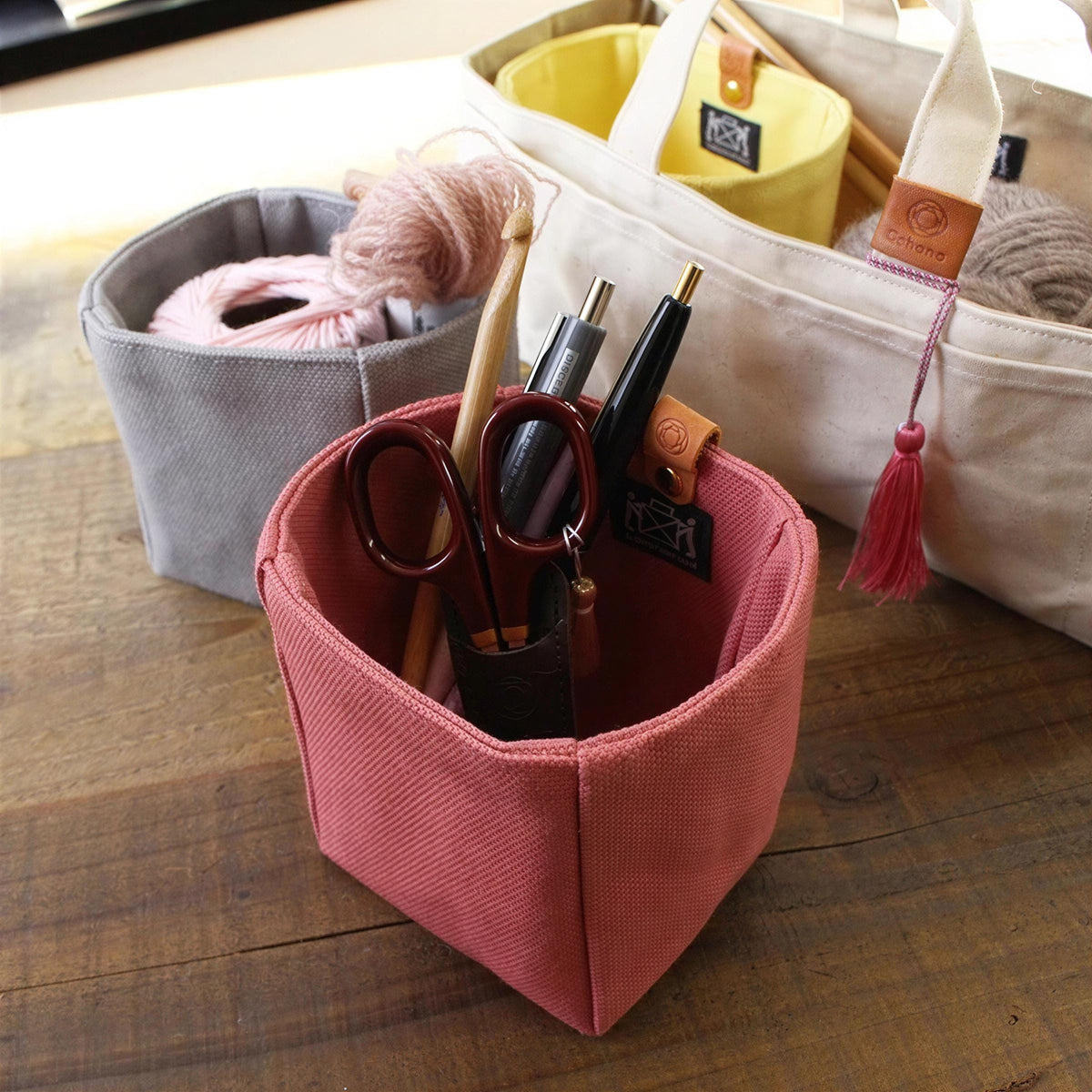 A pink fabric container holding various craft supplies, including scissors, pens, and yarn, sits on a wooden surface.